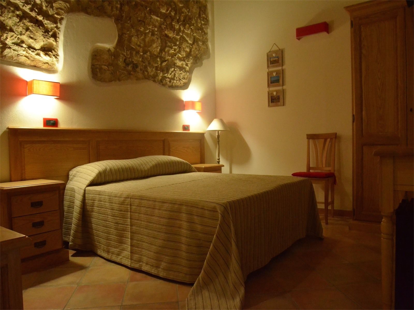 Quarto com cabeceira de madeira, cama com colcha listrada, duas luminárias de cabeceira, mesa de cabeceira com gavetas, uma cadeira e guarda-roupa de madeira.