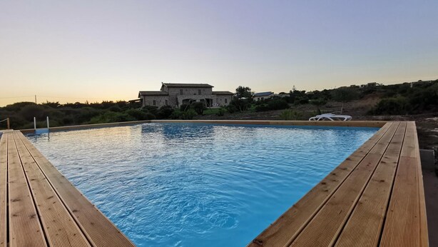 Eine Holzterrasse umgibt einen Swimmingpool mit Blick auf ein Steinhaus im Hintergrund.