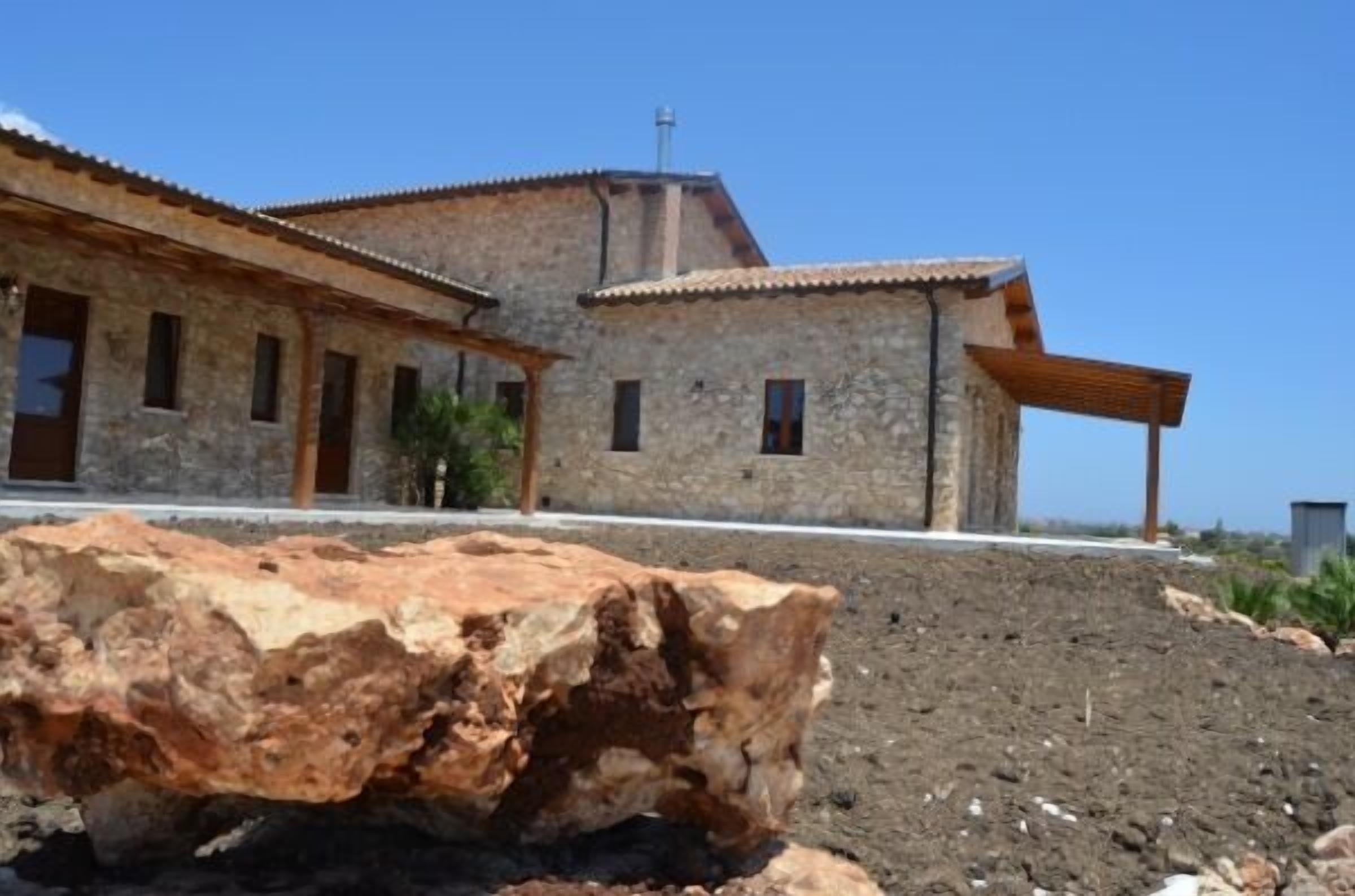 Un bâtiment en pierre avec un toit en tuiles, une pergola en bois et un grand rocher au premier plan.