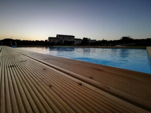Une terrasse en bois mène à une piscine, et on aperçoit une grande maison au loin.