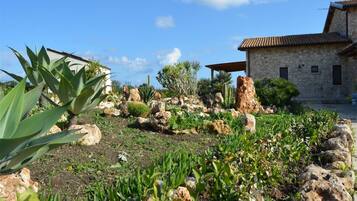 Un bâtiment en pierre, doté d’un toit en tuiles, entouré d’un jardin où poussent différentes plantes et où l’on trouve des rochers.