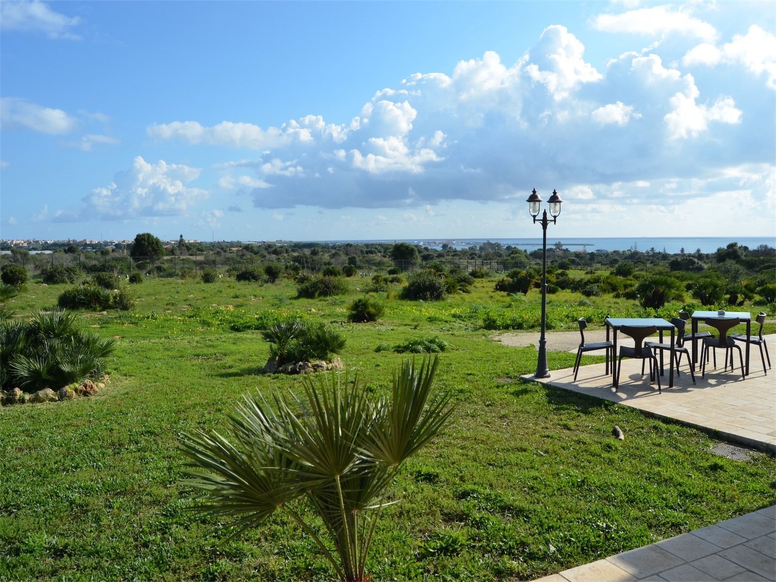 A grassy outdoor area with tables and chairs, a lamp post, and a view of the sea.