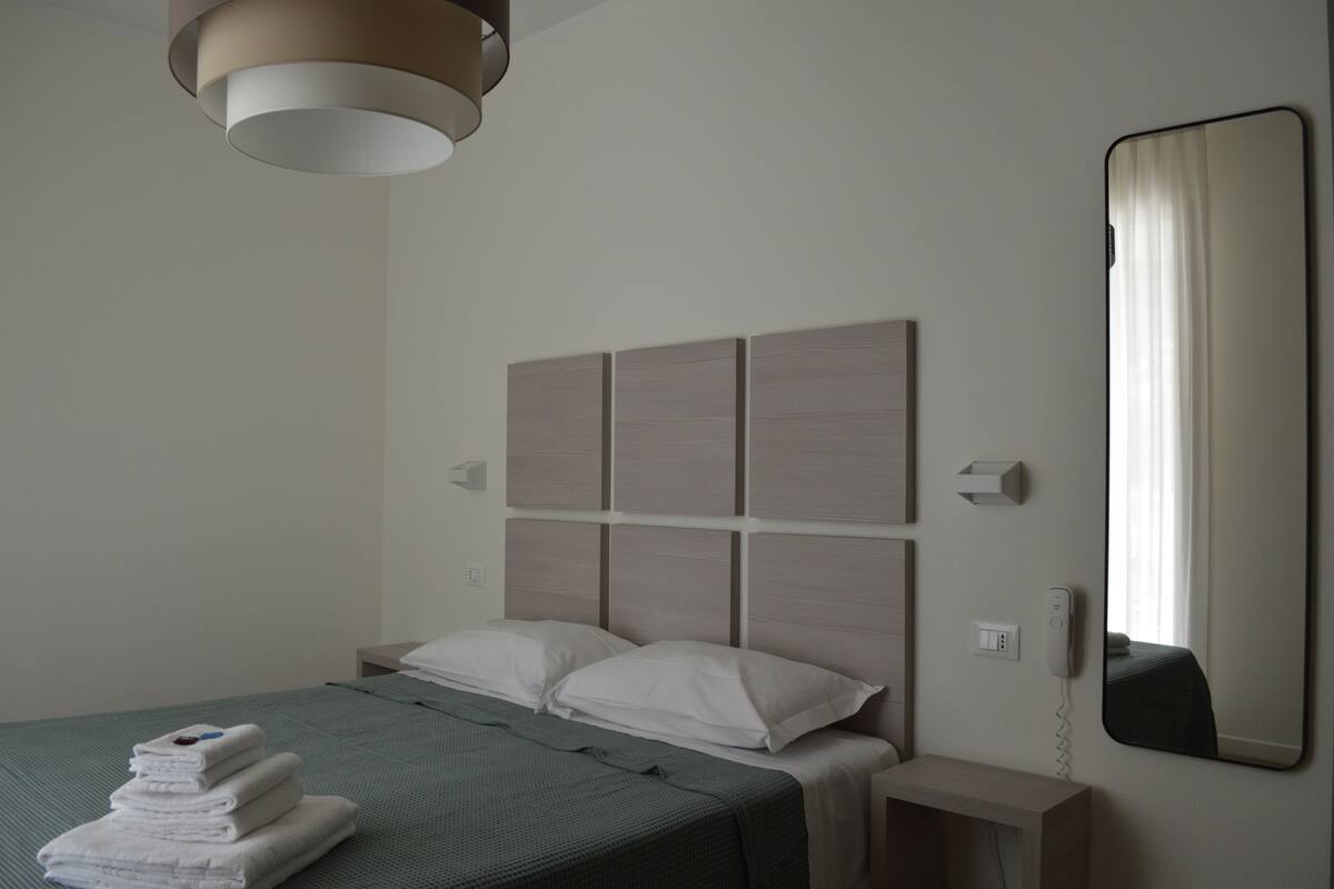 A hotel room with a bed, a bedside table, a wall-mounted telephone, and a large mirror.