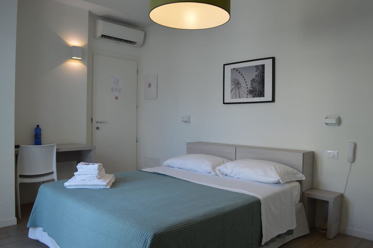 A hotel room with a bed, a desk, a chair, and a wall-mounted air conditioner.