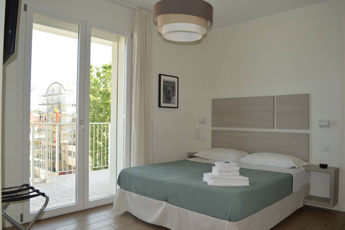 A hotel room with a bed, a bedside table, a chair, a balcony, and a view of the outside.