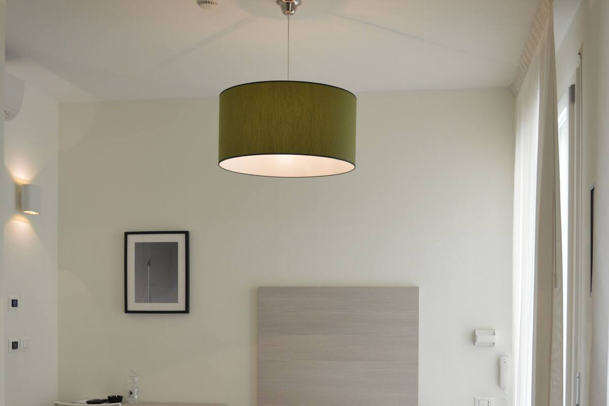 A modern hotel room with a bed, desk, chair, and a hanging light fixture.