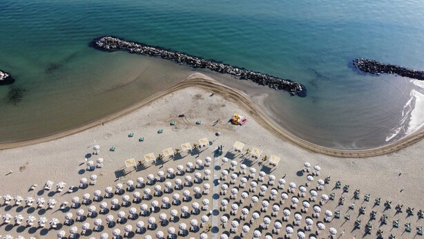 Vista aérea de una playa con hileras de tumbonas y sombrillas, un rompeolas rocoso y agua marina tranquila.