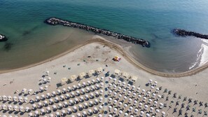 An aerial view of a beach with rows of sunbeds and umbrellas, a rocky breakwater, and calm sea water. - Hotel Riva e Mare (Rimini)
