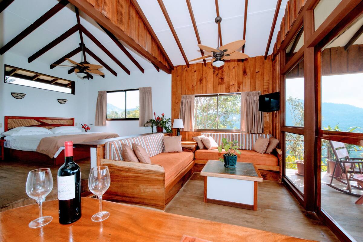 A spacious room with a wooden ceiling, a bed, a sofa, a dining table, and a balcony with a view.