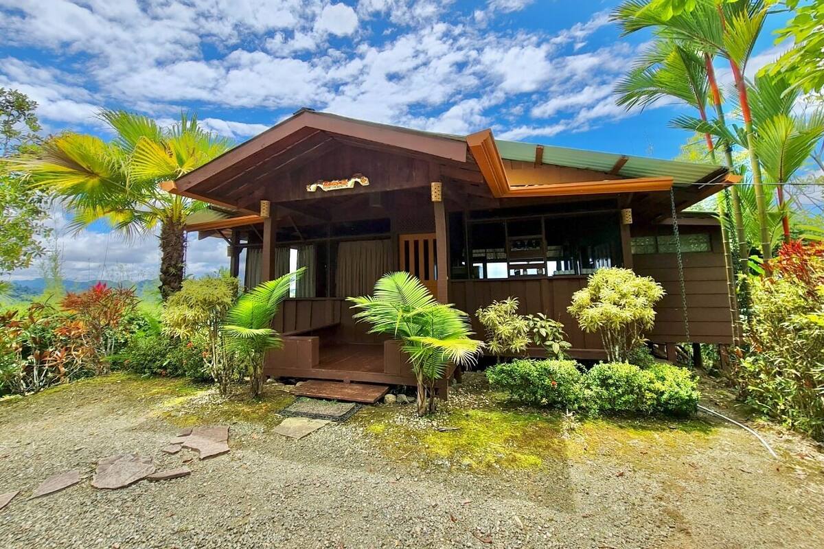 A wooden house with a porch, surrounded by tropical plants and a clear sky.