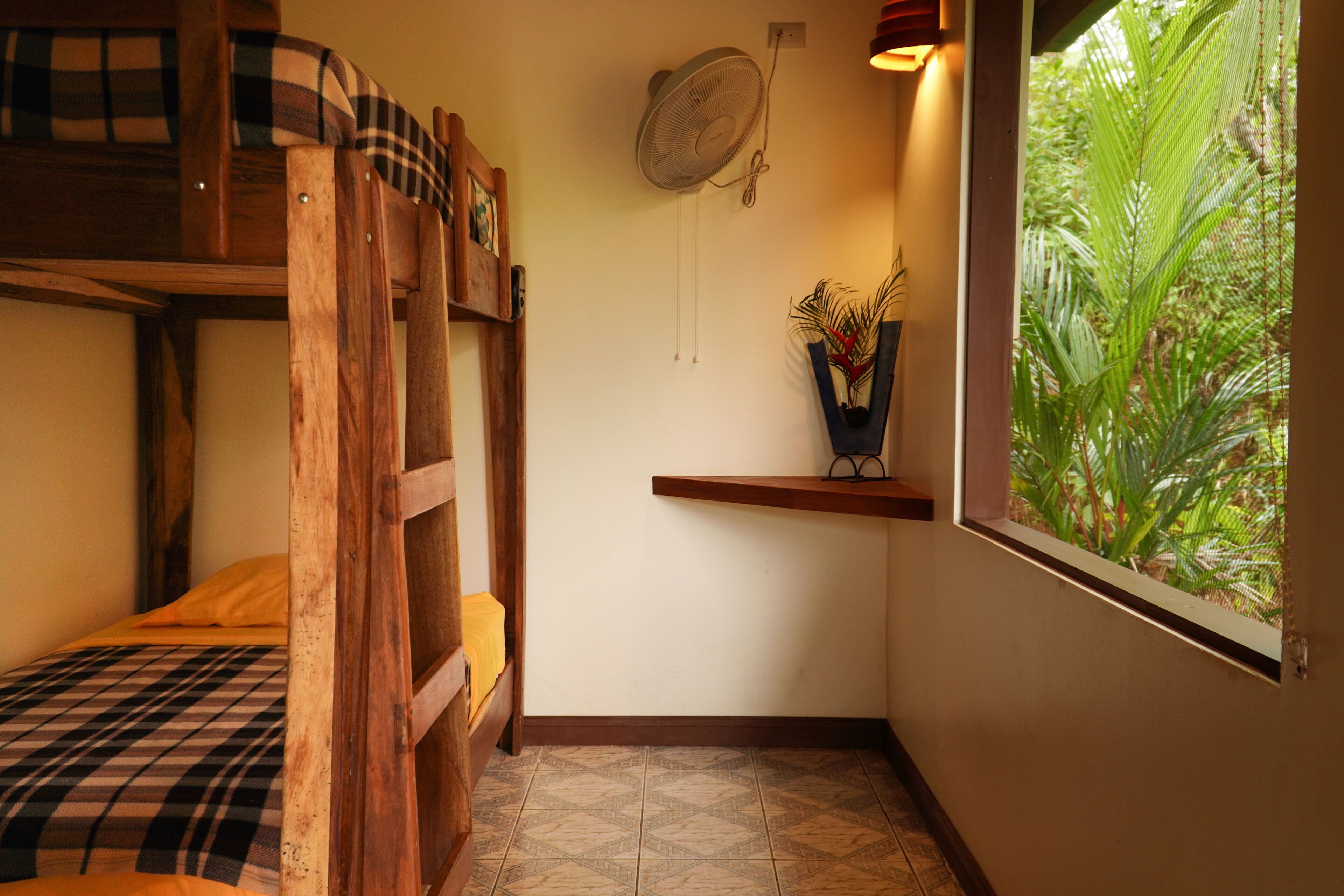 Une chambre d’angle avec un lit superposé, un ventilateur fixé au mur, une étagère avec une plante et une fenêtre donnant sur un paysage verdoyant.