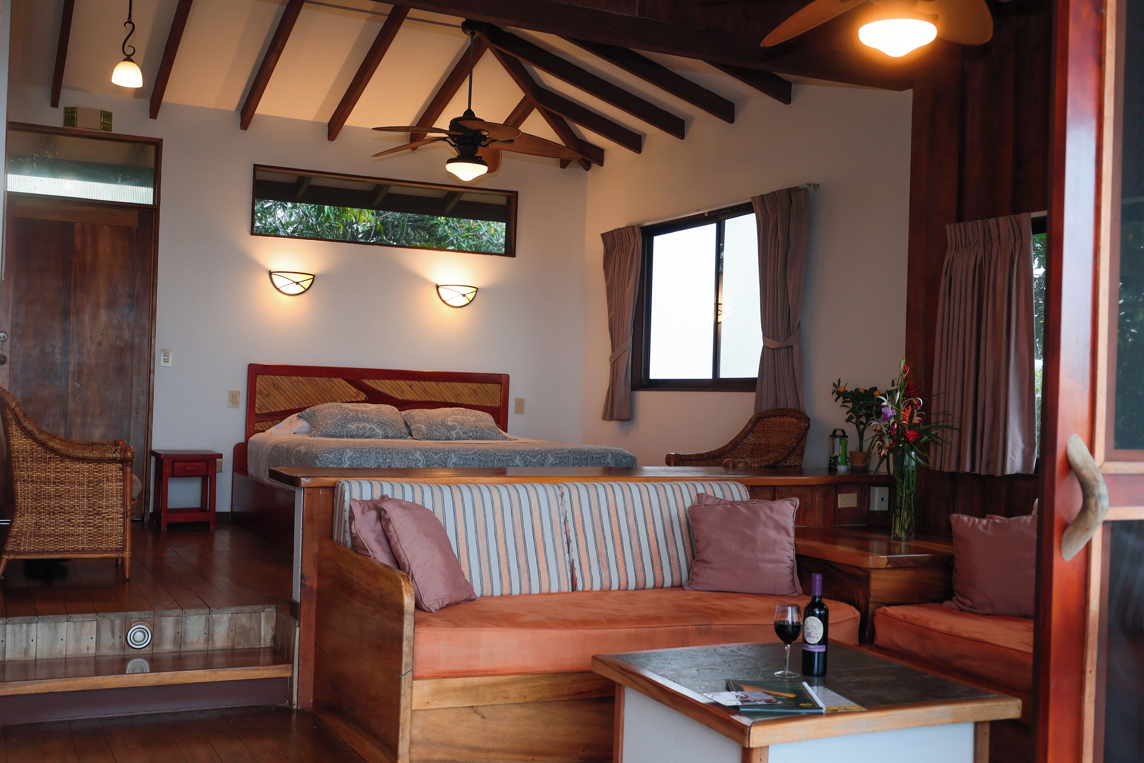 A cozy room with a bed, a striped sofa, a wooden table with a bottle of wine, and a ceiling fan.