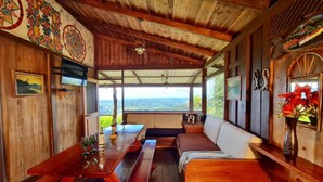 A wooden interior with a wooden bench, a table with a bottle and glasses, and a view of the landscape through large windows. - Santa Juana Lodge and Nature Reserve (Naranjito)