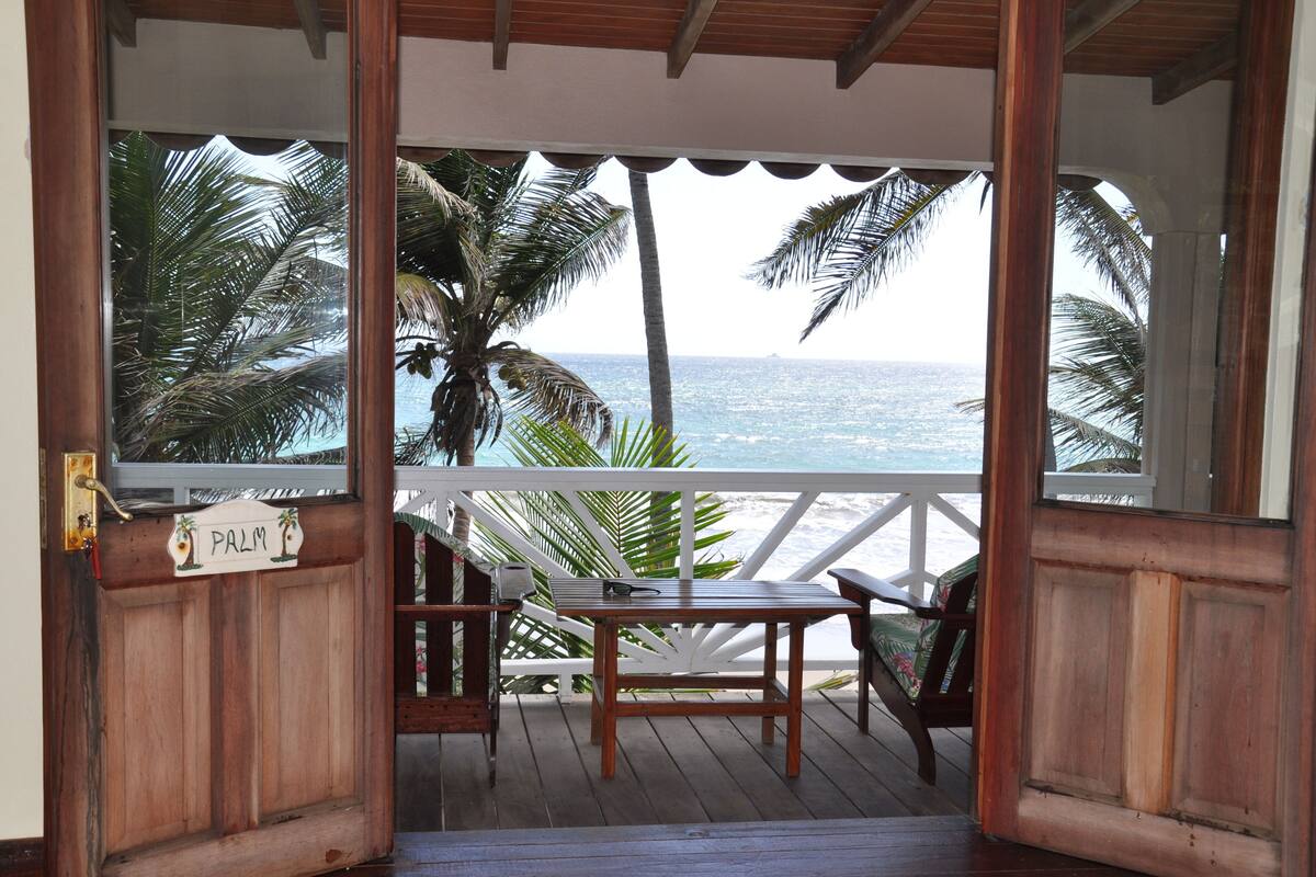 A wooden door with a "PALM" sign, leading to a wooden deck with a table and chairs, overlooking the ocean with palm trees.