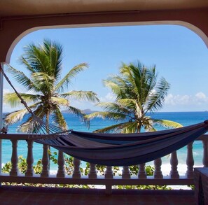 A hammock on a balcony with a view of the ocean and palm trees.