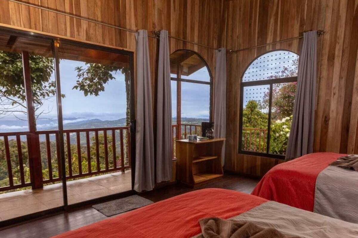 A wooden cabin room with two beds, a desk, and large windows offering a view of the outdoors.