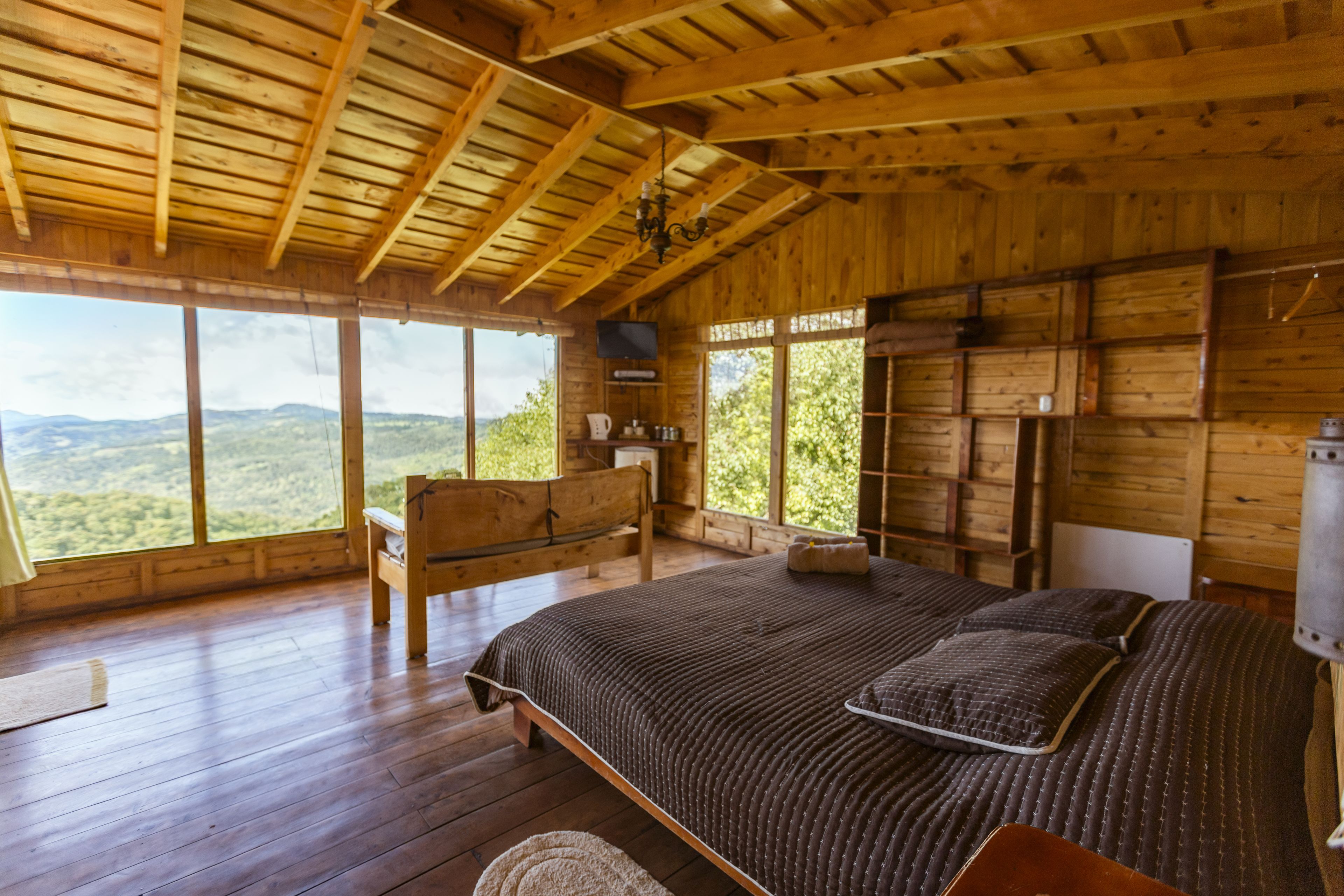 A wooden cabin bedroom with a large bed, a wooden bench, and a view of the outdoors.