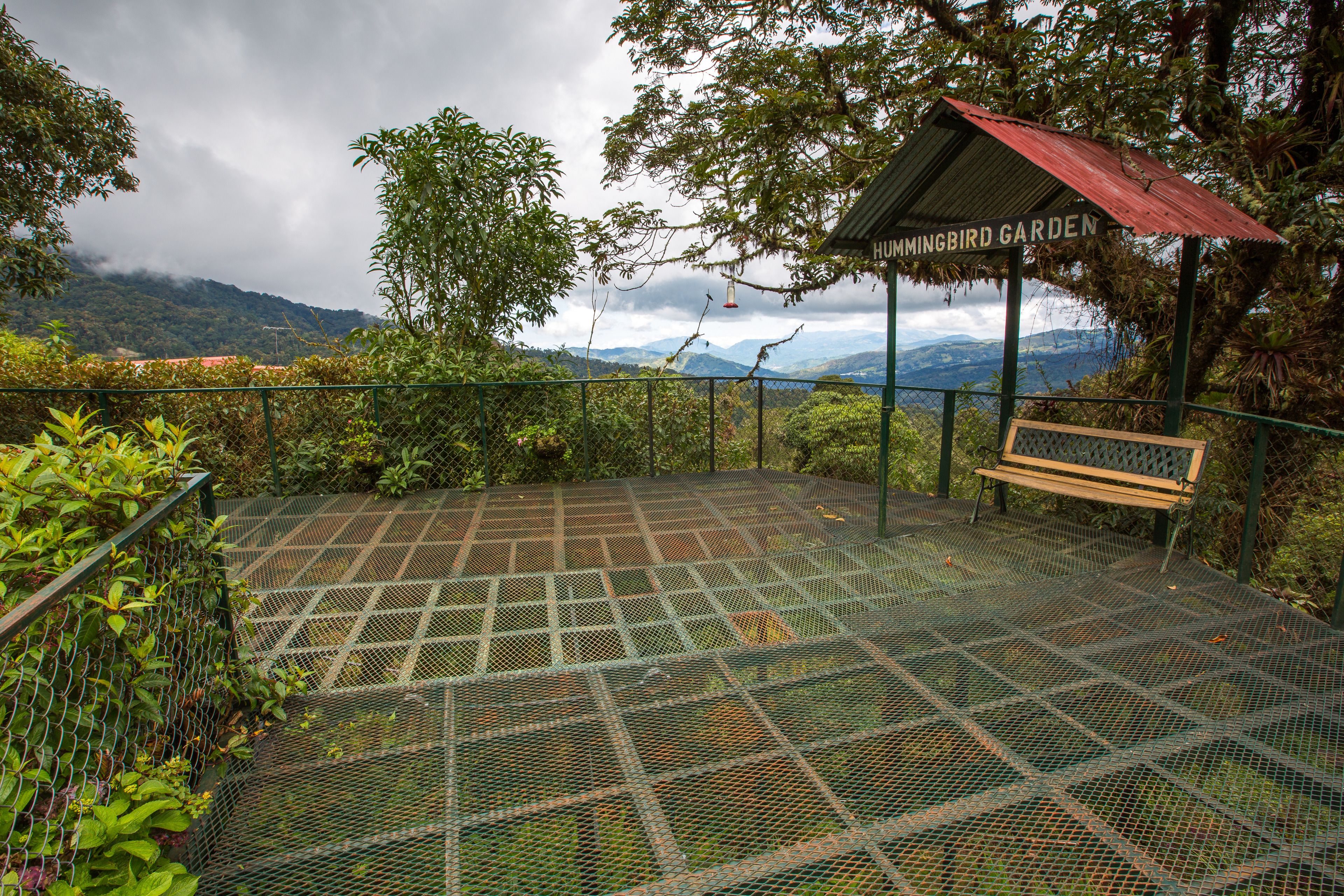 A metal observation deck with a bench, surrounded by a fence and vegetation, overlooking a mountainous landscape.