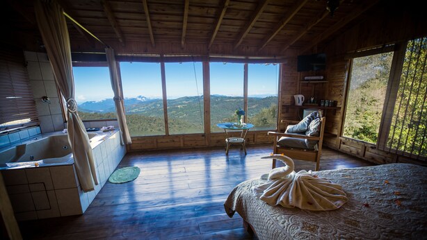A wooden cabin with a large bed, a bathtub, and a view of the mountains. - Paraíso Quetzal Lodge (Copey)