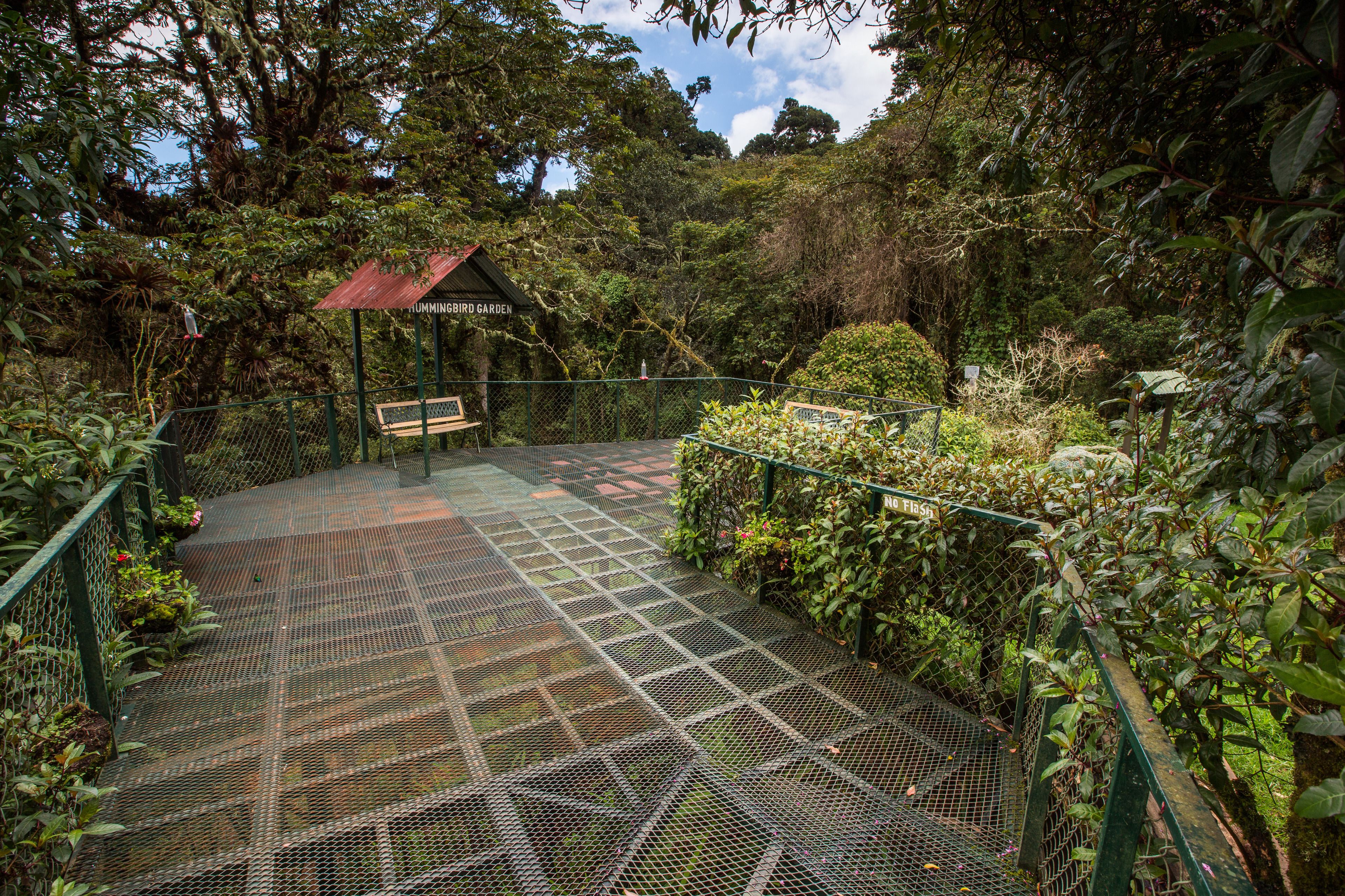 A covered wooden bench in a garden with a metal fence and a sign that reads "Tumblingbird Garden."