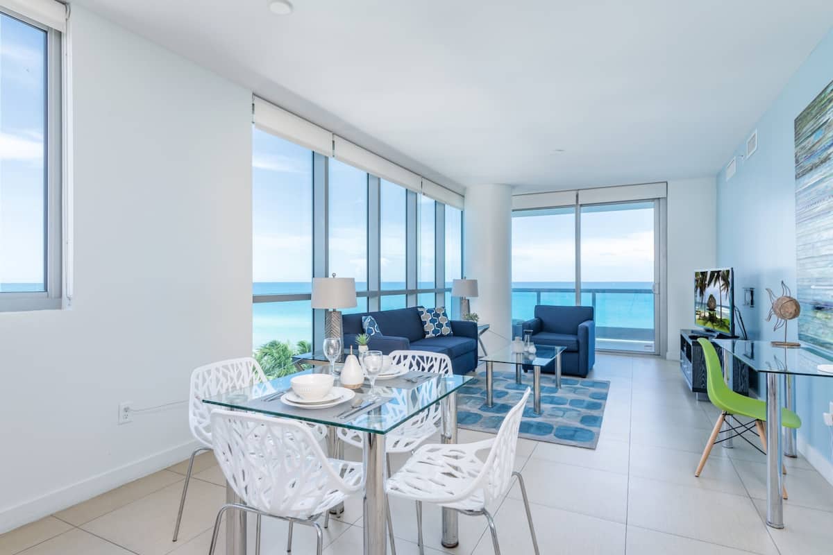 A modern living room with a glass dining table, white chairs, a blue sofa, and a view of the ocean.