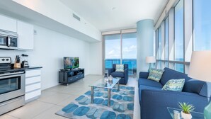 Una sala moderna con un sofá azul, una mesa de centro de vidrio y una zona de cocina con estufa y microondas.