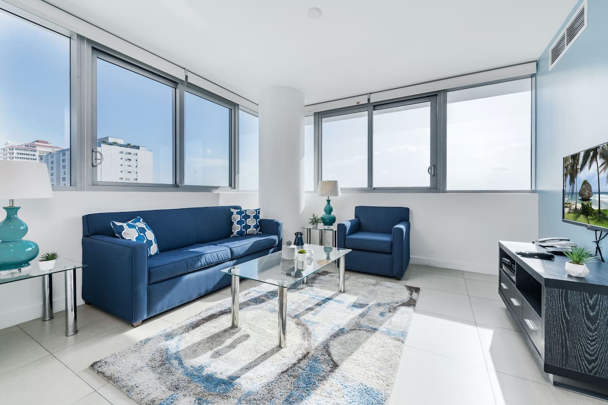 A modern living room with two blue sofas, a glass coffee table, and a large window offering a city view.