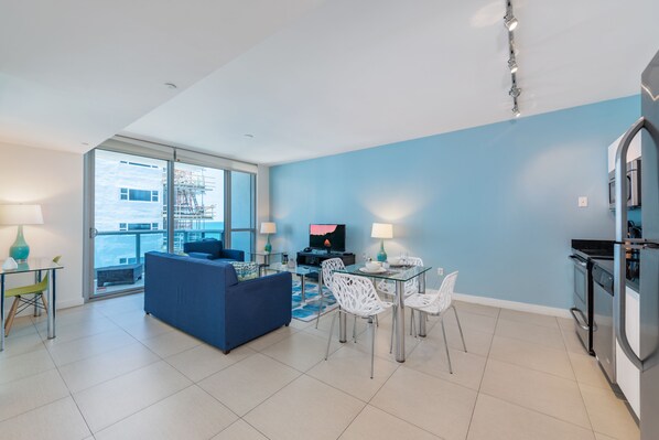 Una sala moderna con un sofá azul, una mesa de comedor de vidrio con sillas blancas, y una zona de cocina con refrigerador y estufa.