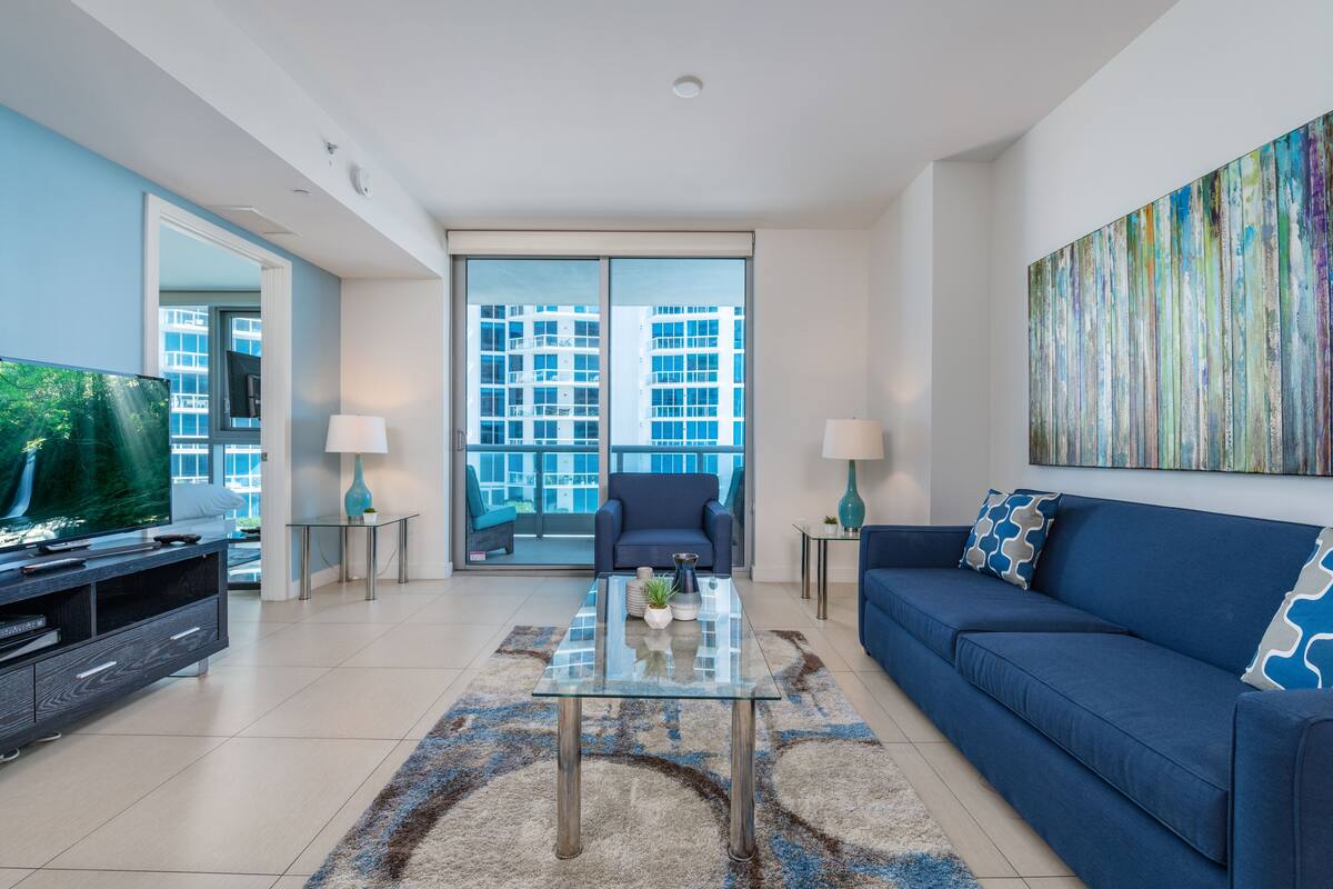 A modern living room with a sofa, a glass coffee table, a TV, and a large window with a view of buildings.
