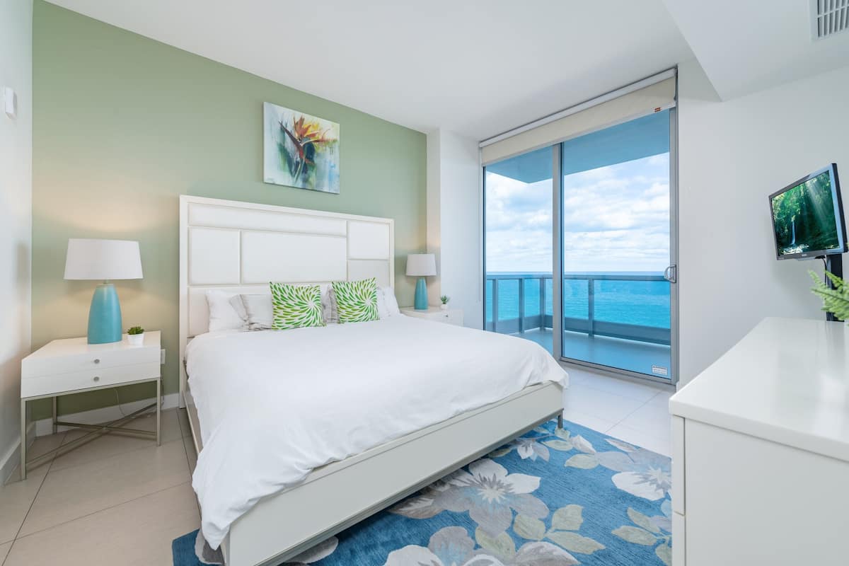 A bedroom with a bed, nightstands, a TV, a painting, and a balcony with a view.