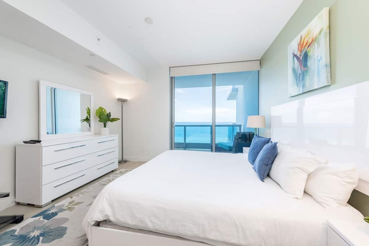 A modern bedroom with a large bed, a dresser, a mirror, and a view of the ocean.