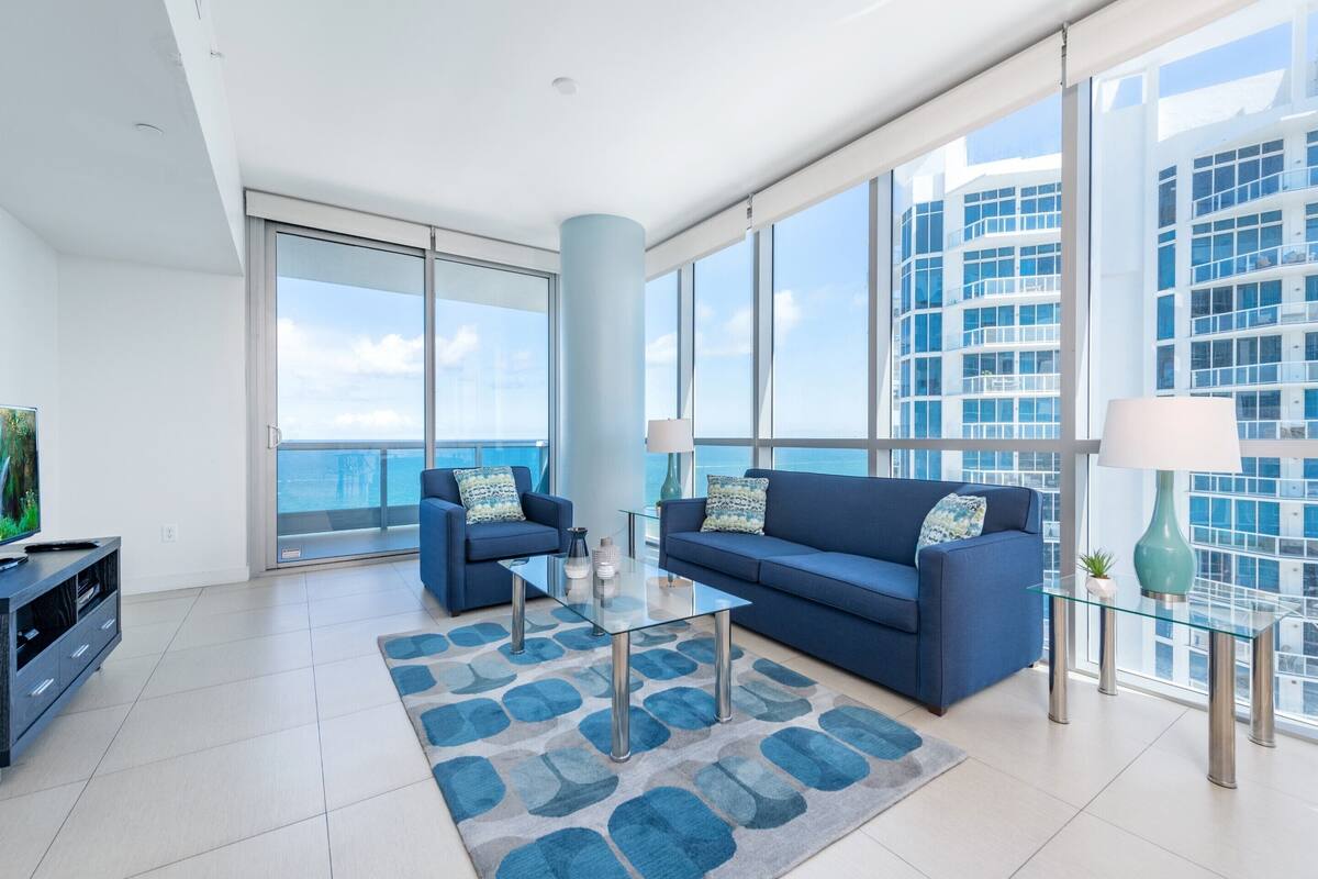 A modern living room with a large glass table, two blue sofas, and a view of the city skyline.