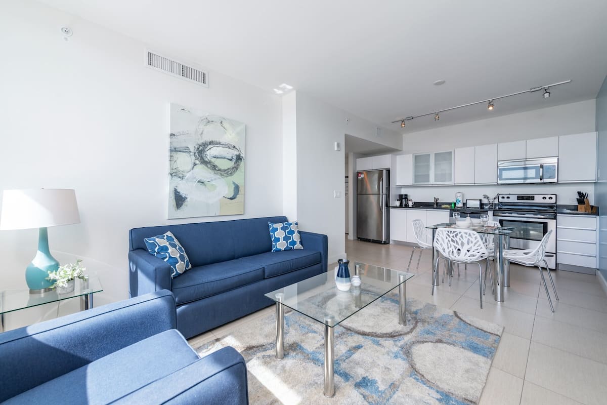 A modern living room with a blue sofa, a glass coffee table, and a dining area with white chairs.