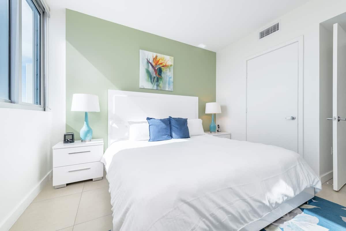 A bedroom with a large bed, white nightstands, a white door, and a window.