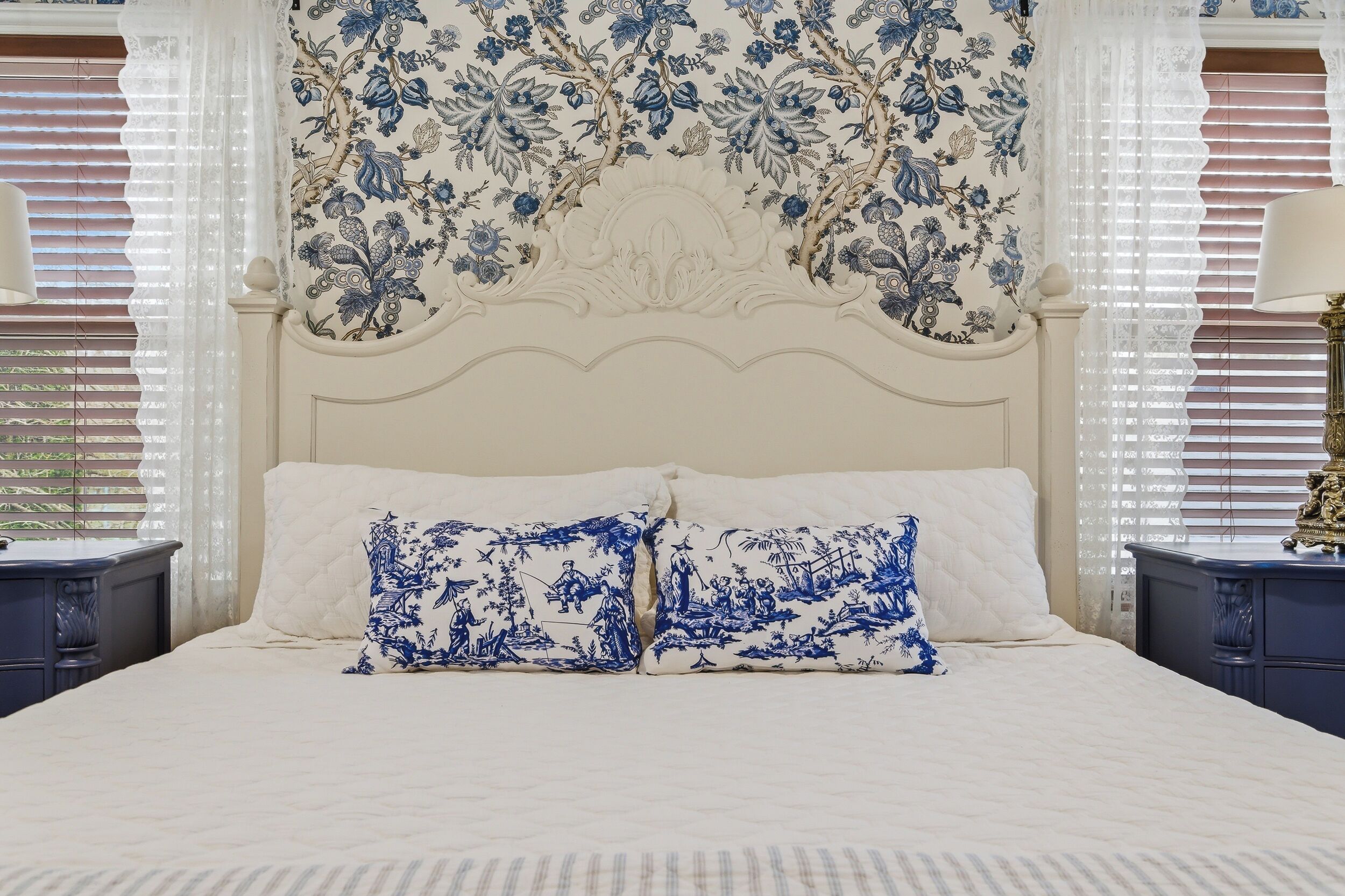 Una cama con ropa de cama blanca y almohadas estampadas con flores azules, un cabecero decorativo y una ventana con persianas.