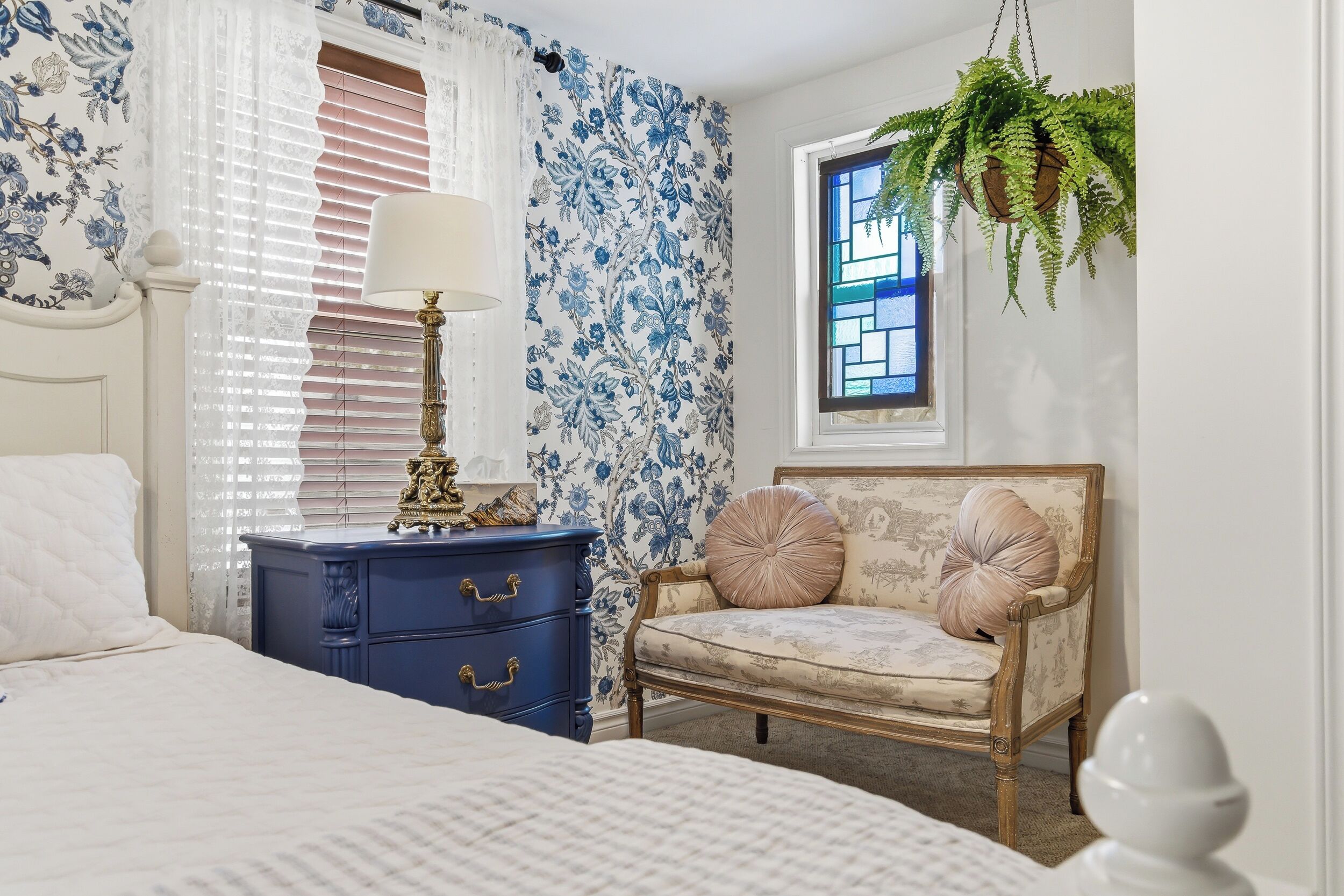 Un dormitorio con cama, cómoda, silla, lámpara, ventana y planta colgante.