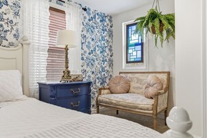 Un dormitorio con cama, cómoda, silla, lámpara, ventana y planta colgante.