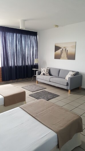 A modern hotel room with a grey sofa, a bed with a beige cover, a small side table, and a painting of a lake. - CIW Hostel (Santa Lucia)