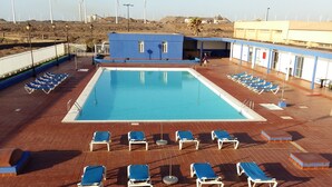 Une piscine entourée de chaises longues bleues, un bâtiment à la façade bleue et blanche, et des éoliennes en arrière-plan.