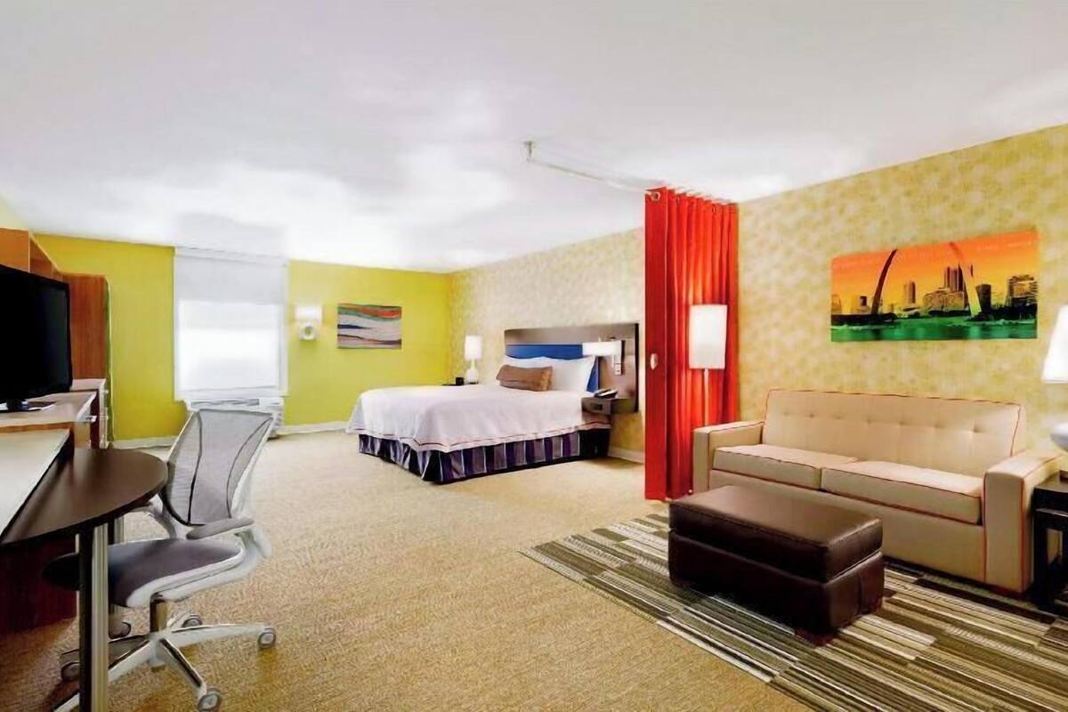 Habitación de hotel con cama, escritorio, silla, sofá, otoman, televisor y un cuadro en la pared.
