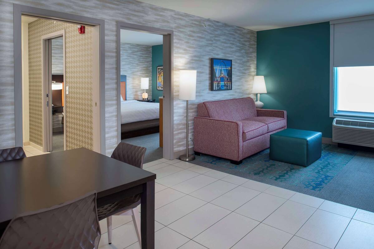Habitación de hotel con cama, sofá rosa, una mesita y una silla. Se ve una ventana con vistas a un edificio.