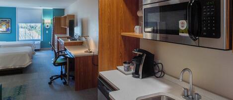 A compact hotel room with a kitchenette, a bed, a desk, and a view of the outdoors.