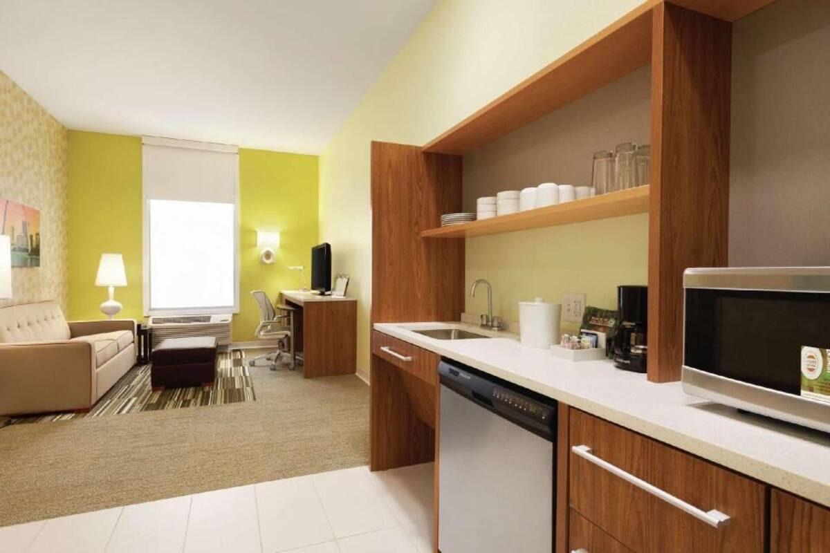 Una habitación de hotel compacta con kitchenette, un sofá, zona de comedor y un ventanal grande.