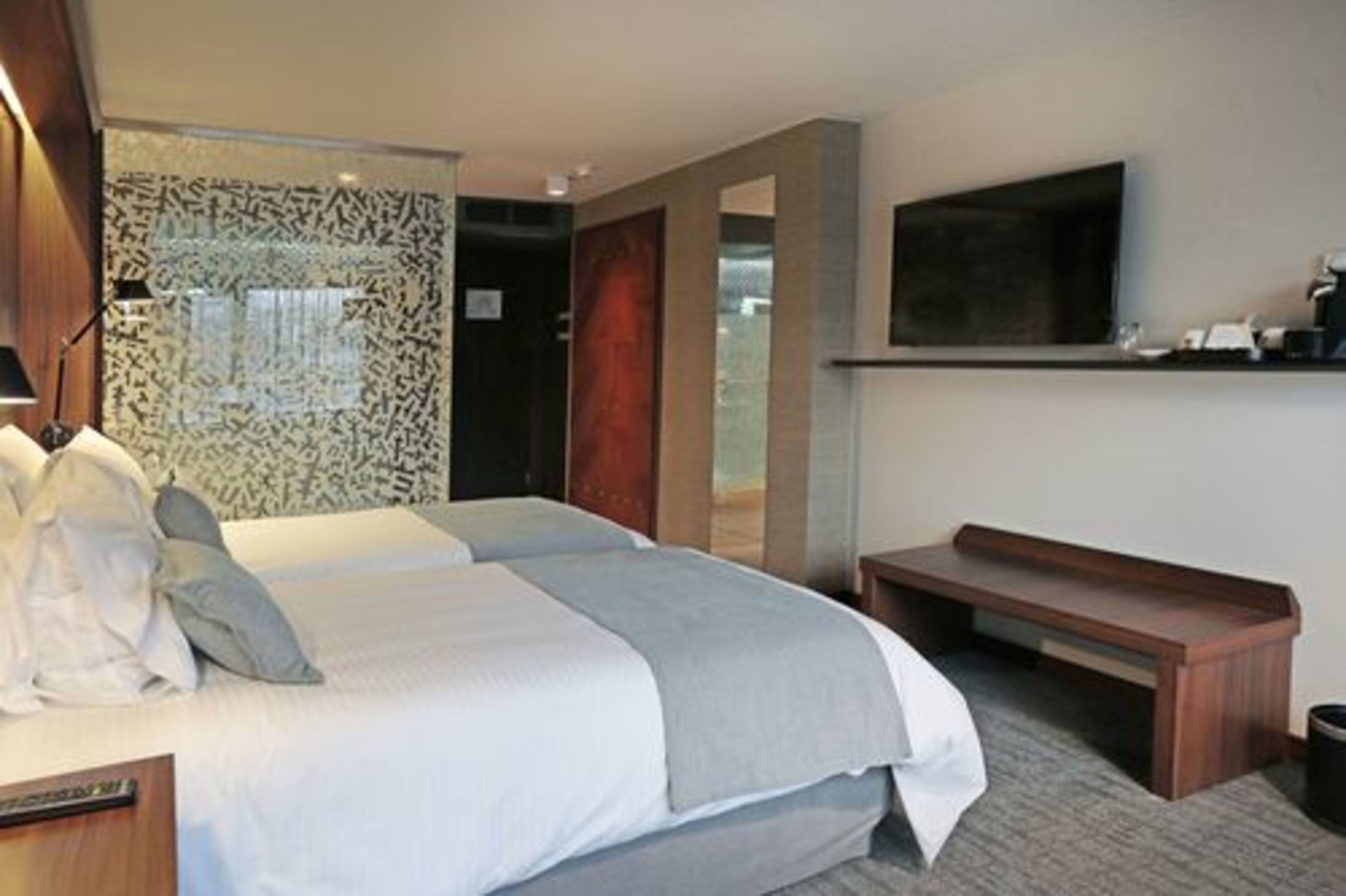 A hotel room with a large bed, a desk, a bench, a flat-screen TV, and a patterned wall.