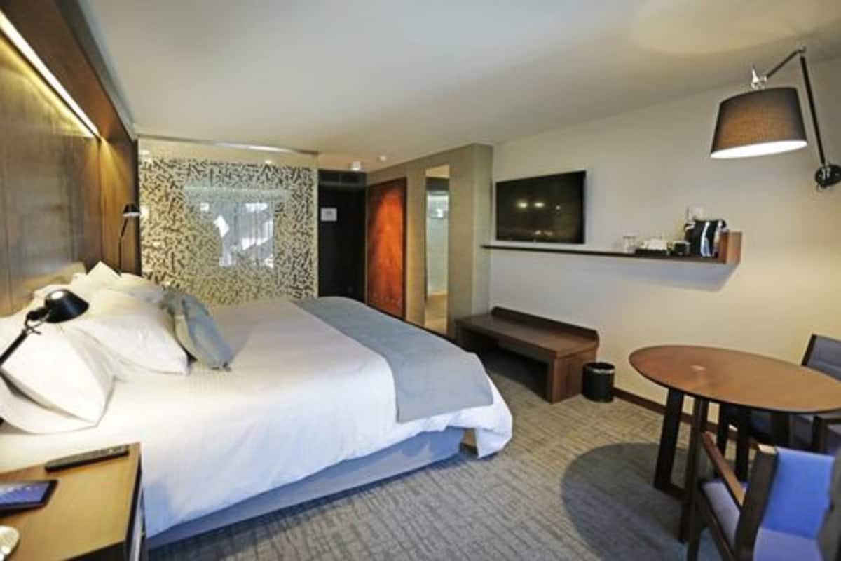 Una habitación de hotel con cama, mesita de noche, lámpara y una pequeña mesa de comedor con sillas.