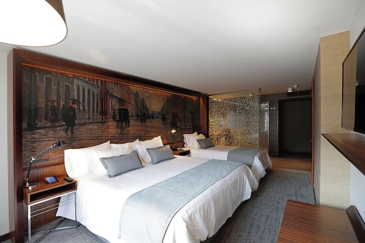 Habitación de hotel con dos camas, un cuadro grande en la pared, un escritorio con lámpara y un televisor a la derecha.