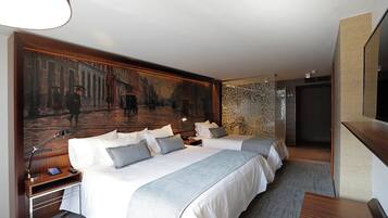 Habitación de hotel con dos camas, un cuadro grande en la pared, un escritorio con lámpara y un televisor a la derecha.