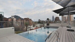 Una piscina al aire libre con cubierta de madera y barandillas de metal, con vistas a un paisaje urbano que incluye edificios y una colina.