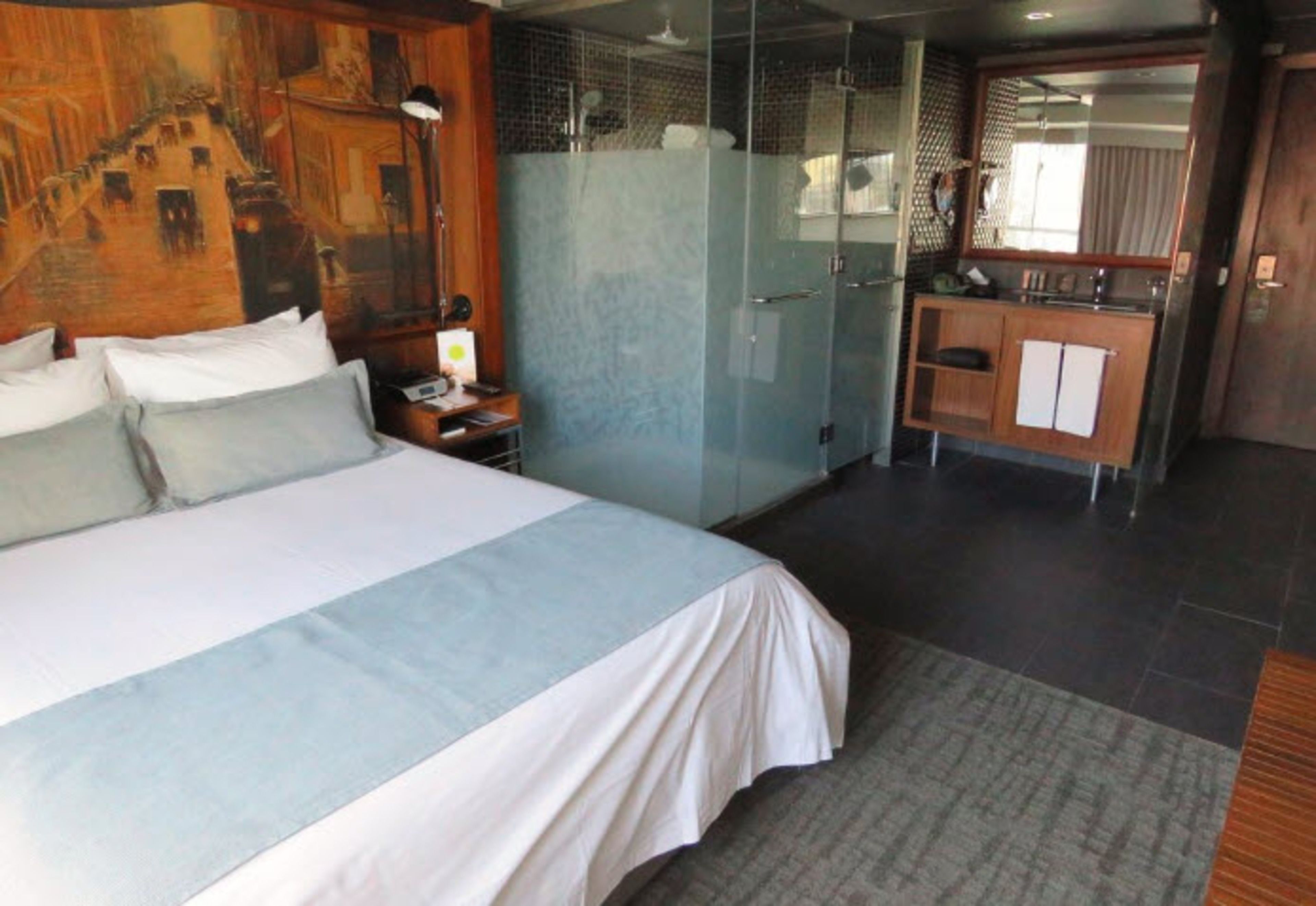 Habitación de hotel con una cama grande, cabina de ducha de vidrio, un escritorio de madera con espejo y un pequeño baño.