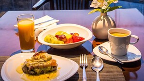 Una mesa con alimentos para el desayuno, un vaso de zumo de naranja y una vista panorámica de un paisaje urbano con edificios y montañas al fondo.